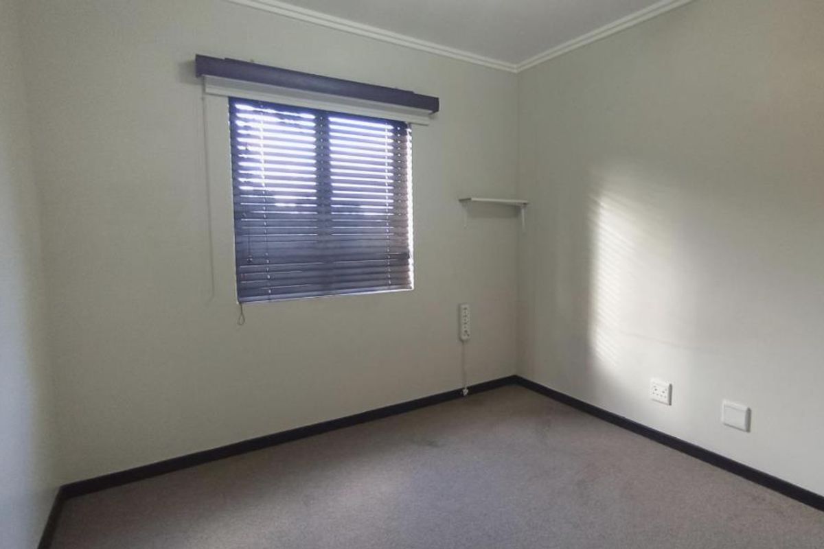 A neutral, empty bedroom featuring carpeted flooring, a window with venetian blinds, and wall-mounted electrical points.