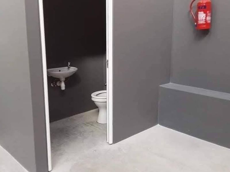 A clean, minimalist industrial-style bathroom featuring a wall-mounted basin, toilet, and a wall-mounted fire extinguisher in a neutral grey finish.