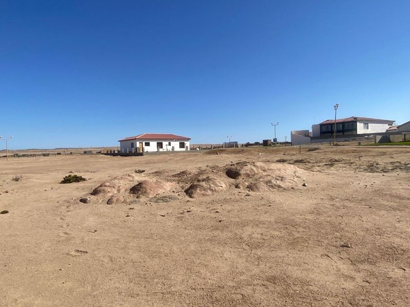 A wide-angle view of a vacant residential plot featuring natural terrain and rock formations, situated in a developed coastal neighborhood.