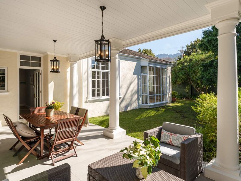 A well-maintained covered patio featuring classical columns, beadboard ceiling, and pendant lighting, overlooking a lush garden with a bay window view.