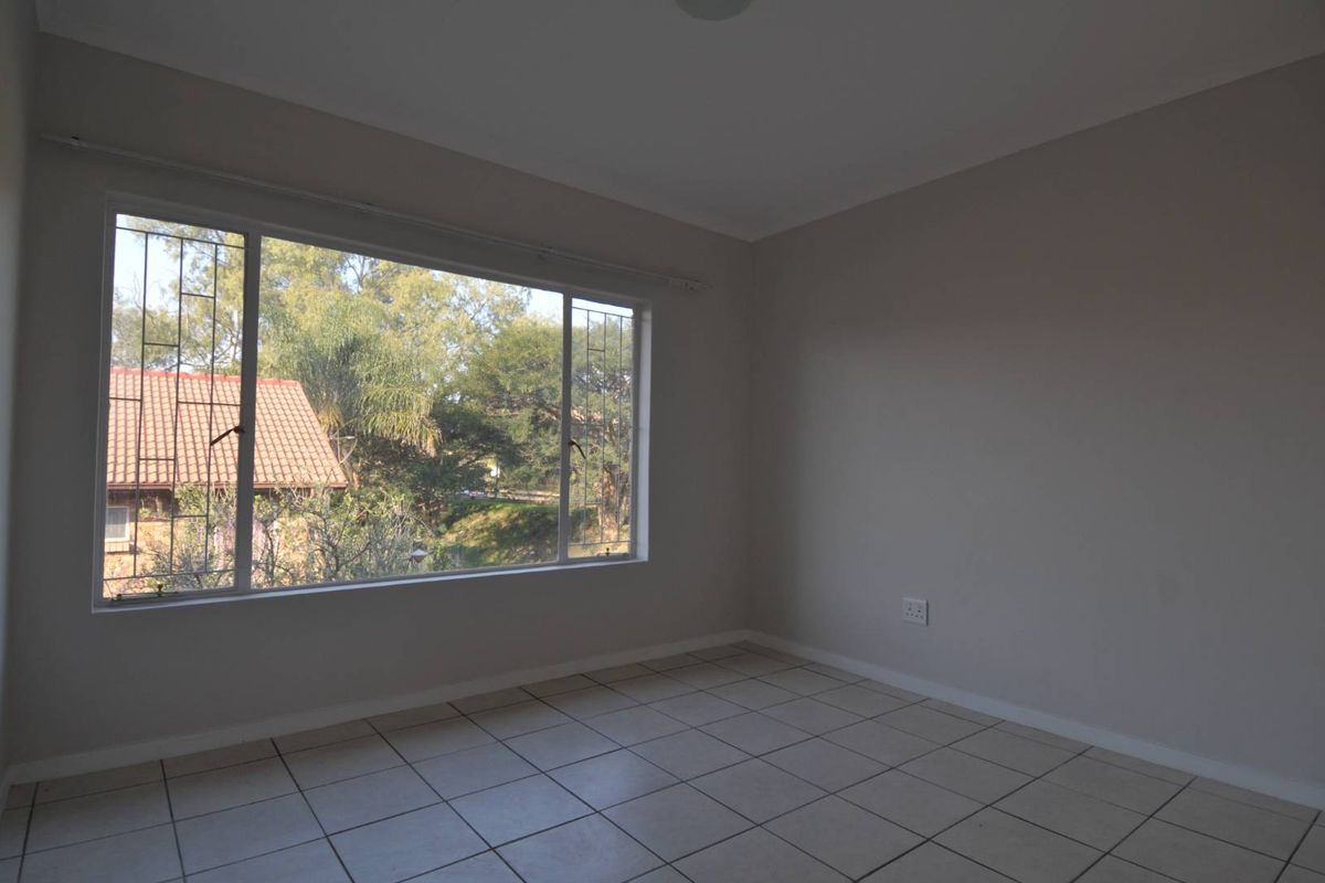 A bright, empty bedroom featuring neutral wall tones, tiled flooring, and a large window with security bars overlooking the surrounding greenery.