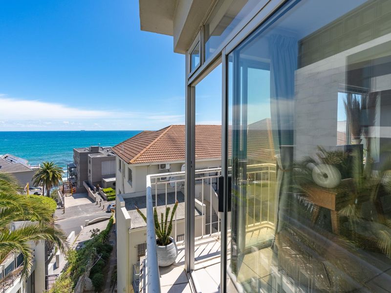 A bright balcony featuring a metal balustrade and tiled flooring, offering expansive coastal and neighbourhood views through large sliding glass doors.