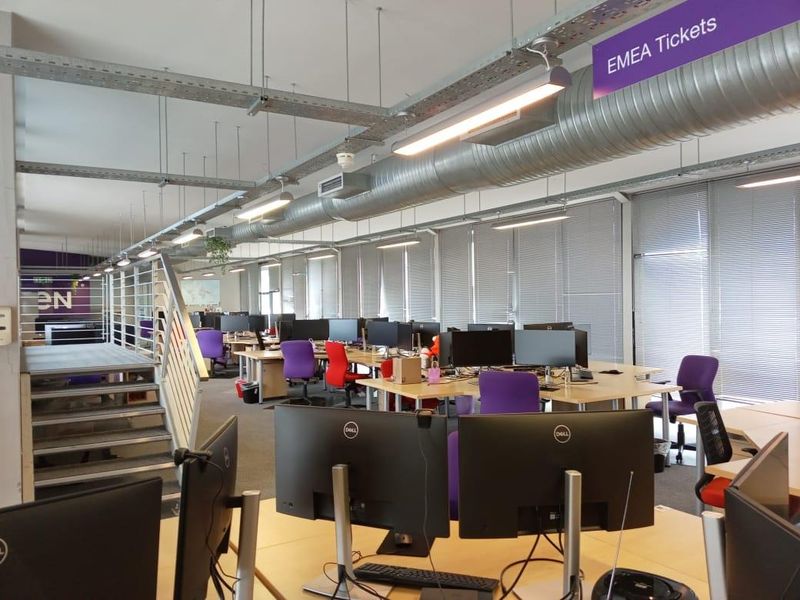 Open-plan office space featuring exposed industrial ductwork, suspended lighting, and large windows with venetian blinds.