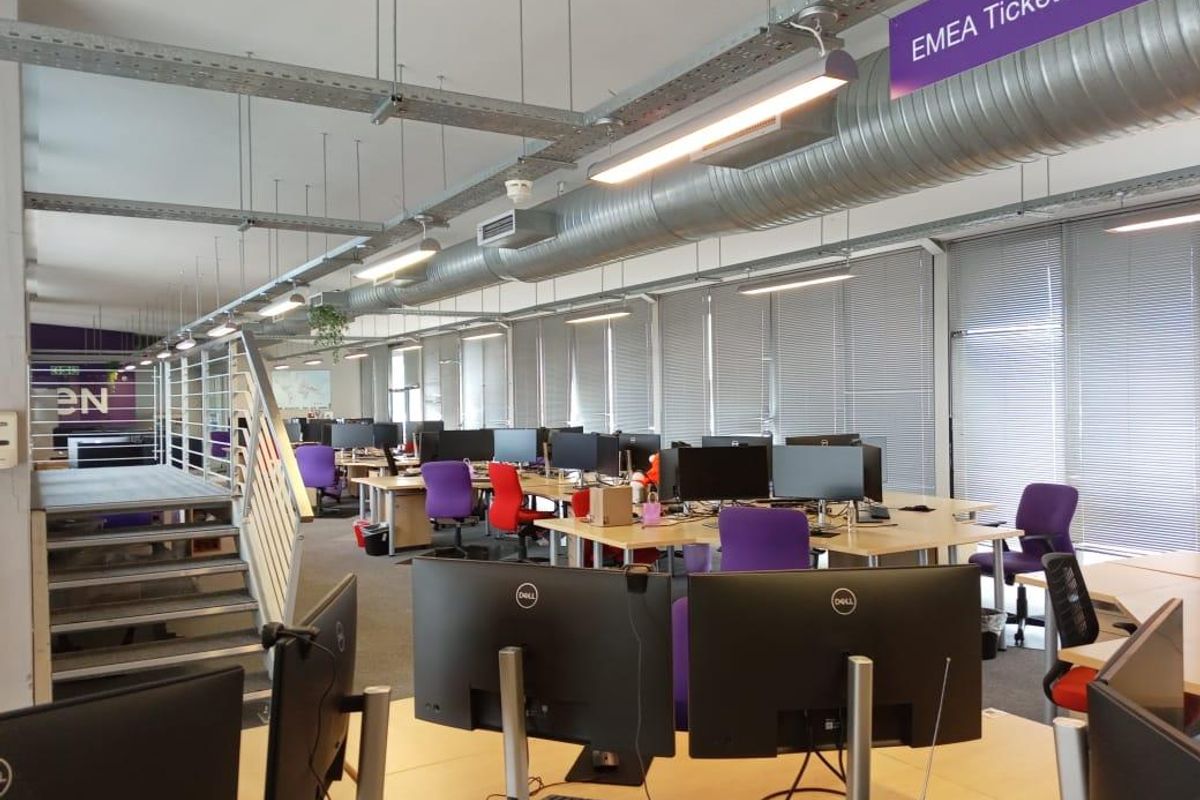Open-plan office space featuring exposed industrial ductwork, suspended lighting, and large windows with venetian blinds.