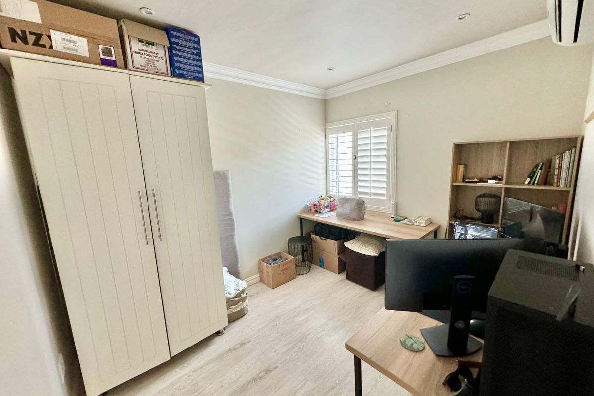 A functional bedroom featuring light laminate flooring, plantation shutters, and an integrated air conditioning unit, currently utilized as a home office space.