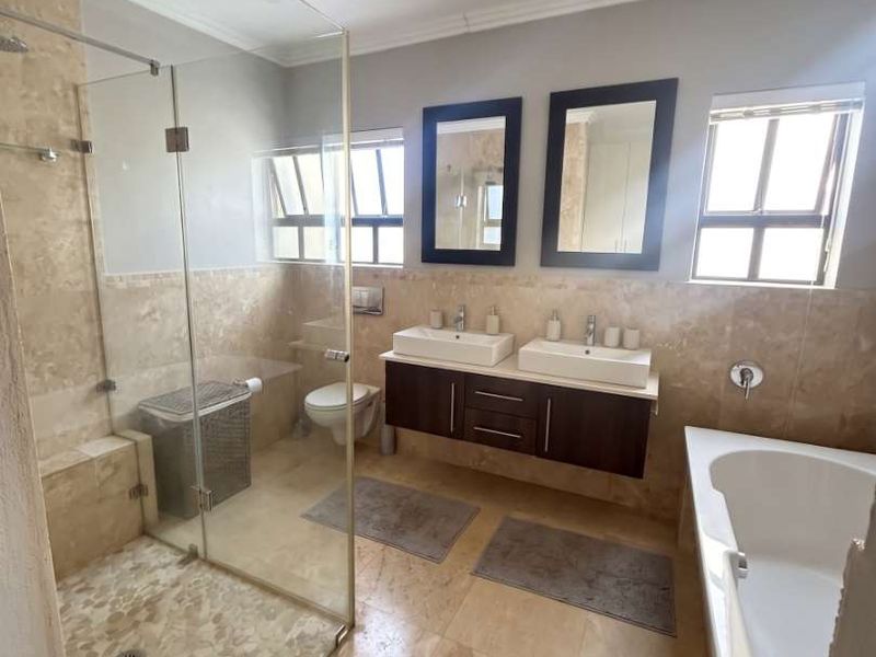 Modern bathroom featuring a double vanity, glass-enclosed shower, deep soaking bathtub, and neutral stone tiling throughout.
