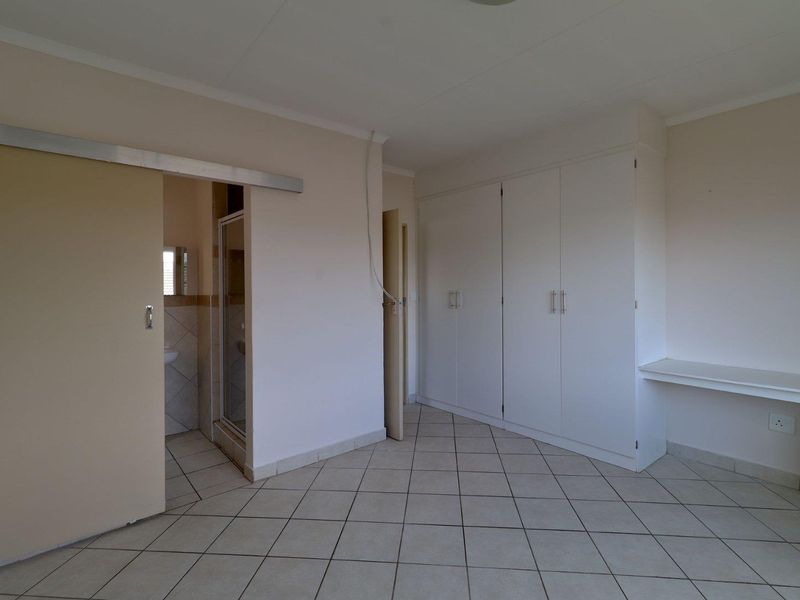 A spacious bedroom featuring neutral tiled flooring, ample built-in cupboard storage, a built-in desk, and direct access to an en-suite bathroom.