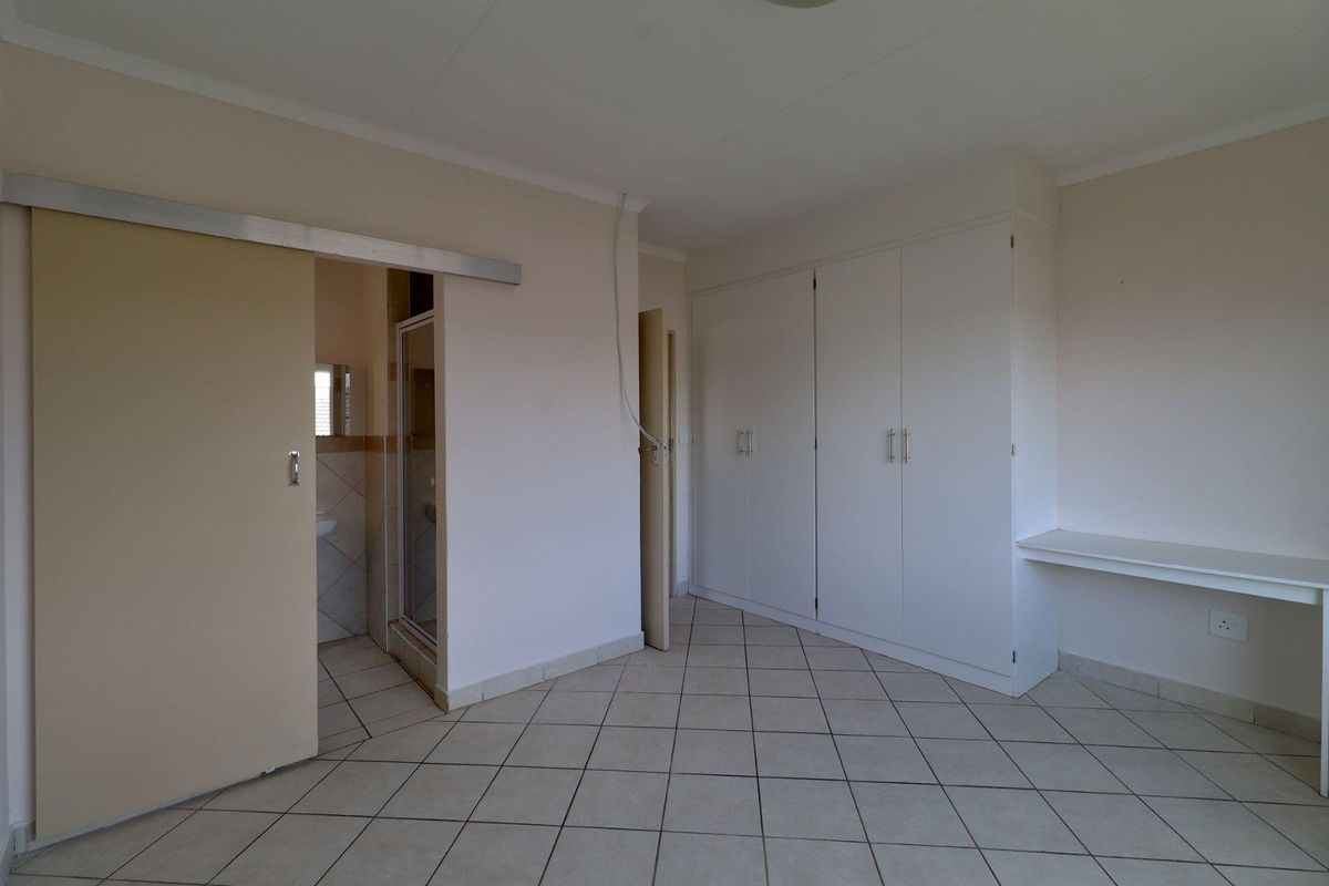 A spacious bedroom featuring neutral tiled flooring, ample built-in cupboard storage, a built-in desk, and direct access to an en-suite bathroom.