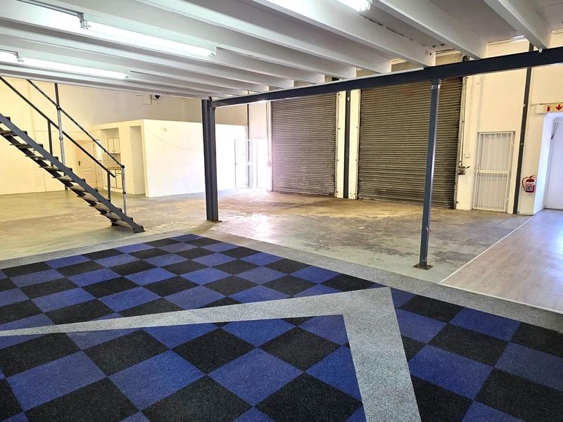 Spacious industrial open-plan unit featuring exposed ceiling beams, a metal staircase, roller shutter doors, and a mix of tiled and concrete flooring.