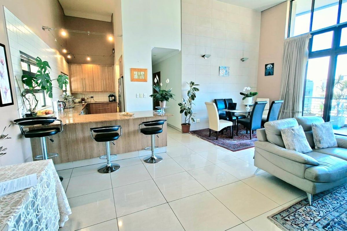 Spacious open-plan living area featuring a modern kitchen with breakfast bar, dining space, and high ceilings with large windows leading to a balcony.