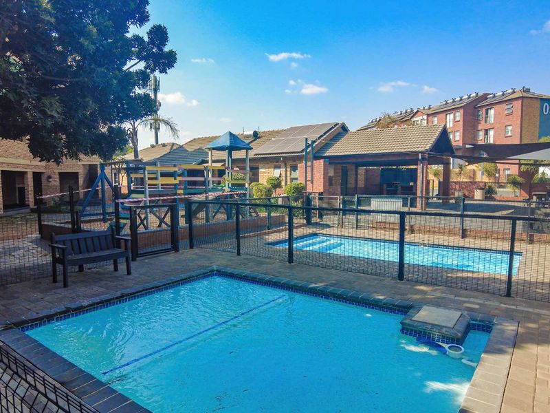 Communal outdoor area featuring a swimming pool, paved surrounds, safety fencing, and a children's playground set within an apartment complex.