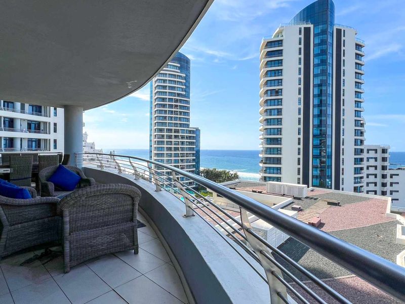 Spacious covered balcony featuring tiled flooring and a stainless steel railing, offering panoramic ocean and city skyline views.
