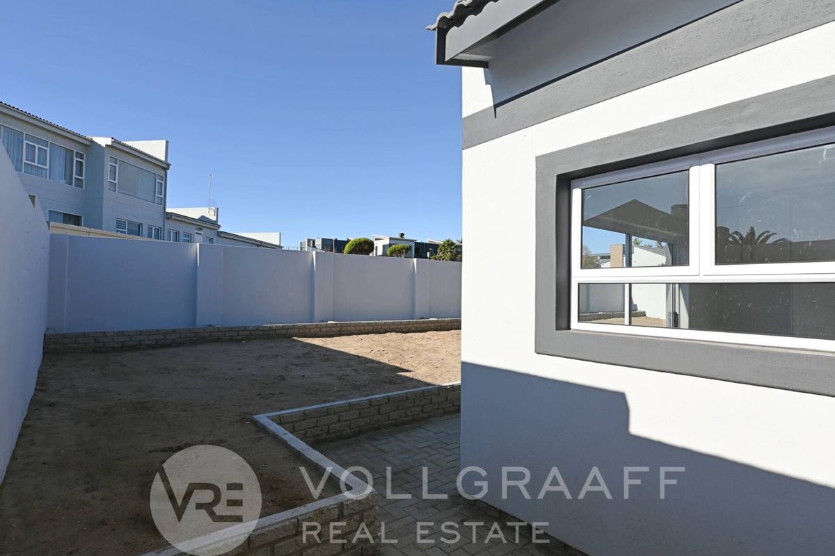 A rear exterior view showing a paved walkway, retaining wall, and an unlandscaped garden area enclosed by a high boundary wall.