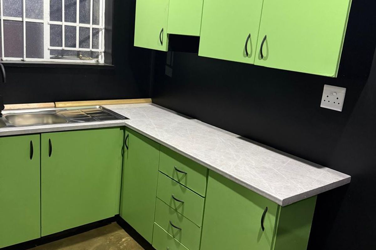 A compact kitchen featuring bright green cabinetry, light-toned laminate countertops, a stainless steel sink, and polished concrete flooring against a bold black wall.