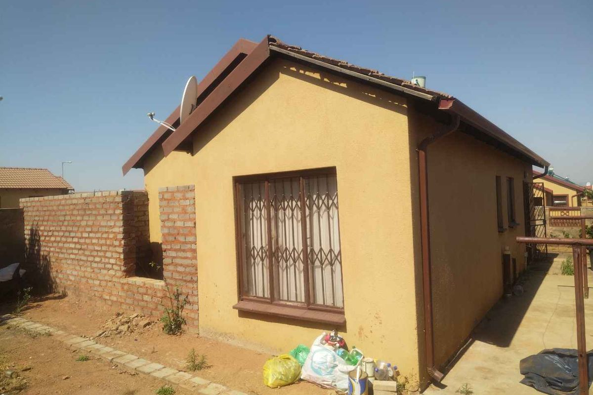 Rear view of a detached house featuring a tiled roof, security gate, and unfinished brick boundary wall in a residential setting.