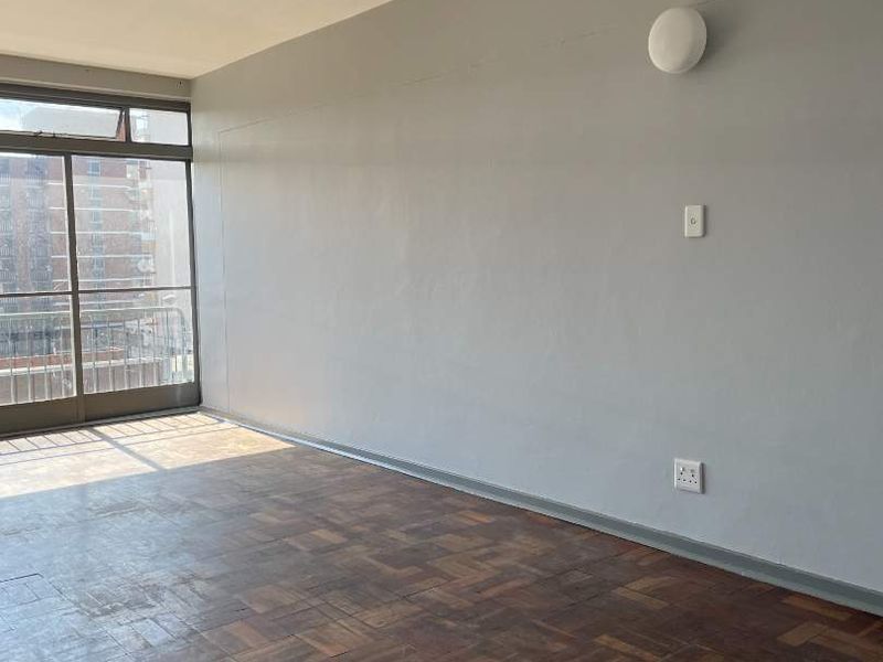 An empty living room featuring classic parquet flooring, neutral grey walls, and a sliding glass door providing access to a balcony.