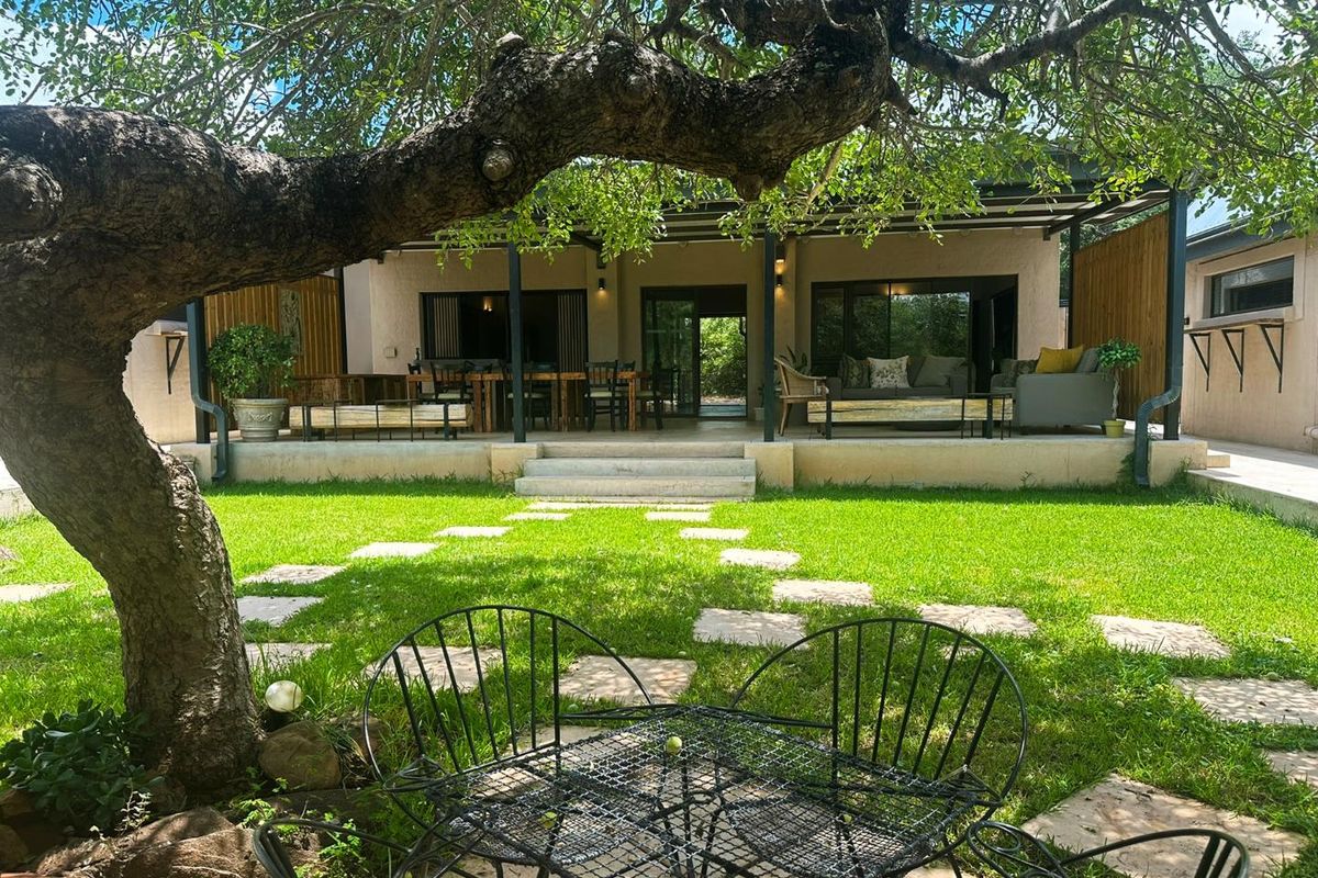 A spacious covered patio overlooking a lush green garden, featuring stone pavers and a large mature tree providing natural shade.