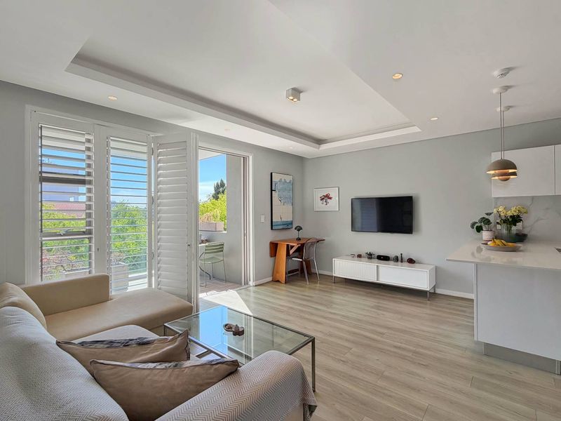 Modern open-plan living area featuring laminate flooring, recessed ceiling with downlights, and a sleek kitchen island, seamlessly connecting to a private balcony.