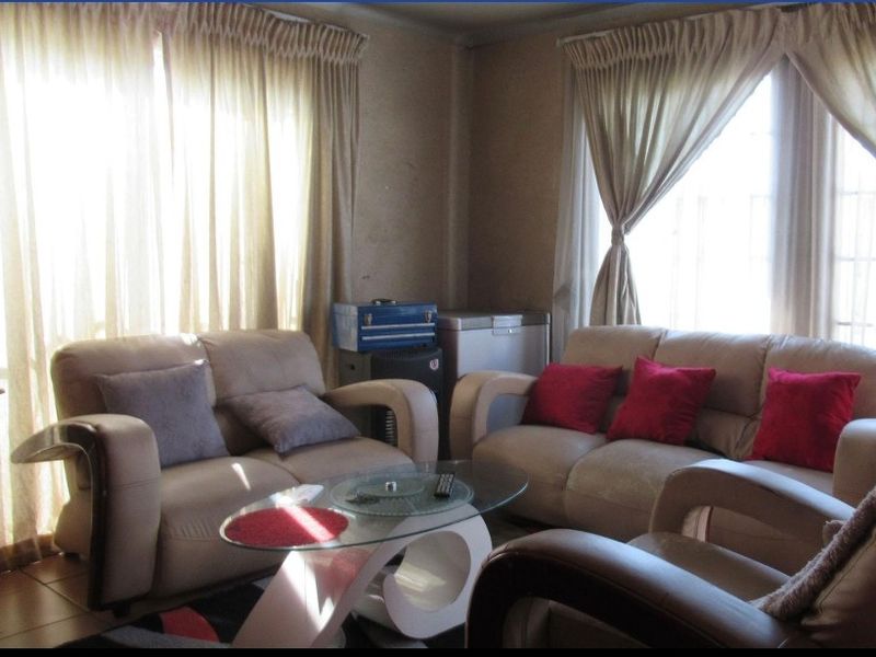 A living room area featuring tiled flooring and large windows with curtain rails, currently showing signs of wear on the wall surfaces.