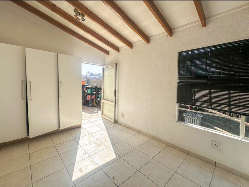 A bright, empty bedroom featuring exposed timber ceiling beams, tiled flooring, built-in cupboards, and direct access to an outdoor patio area.