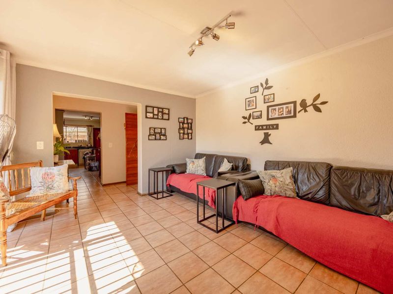 A spacious living room featuring neutral walls, tiled flooring, and track lighting, with an open-plan flow leading into the adjacent kitchen area.