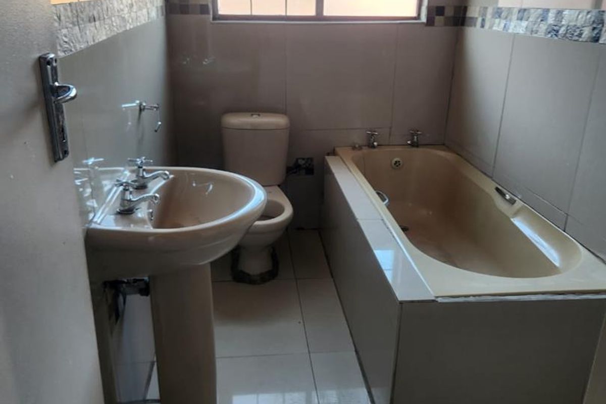 A functional bathroom featuring a tiled finish, a pedestal basin, a toilet, and a built-in bathtub with a window for natural light.