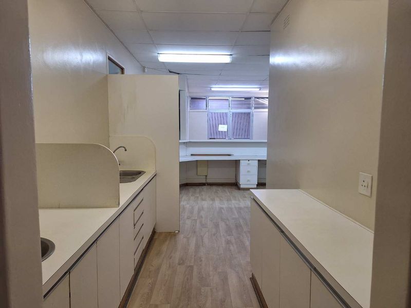 An empty office space featuring built-in cabinetry, a kitchenette area with a sink, laminate flooring, and functional fluorescent lighting.
