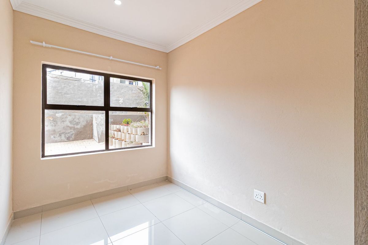 A bright, empty bedroom featuring neutral wall tones, tiled flooring, and a large aluminium-framed window overlooking the rear exterior area.