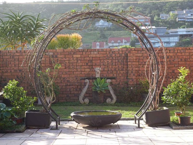 A landscaped garden area featuring a circular metal arch, stone water feature, and brick boundary wall with paved patio flooring.