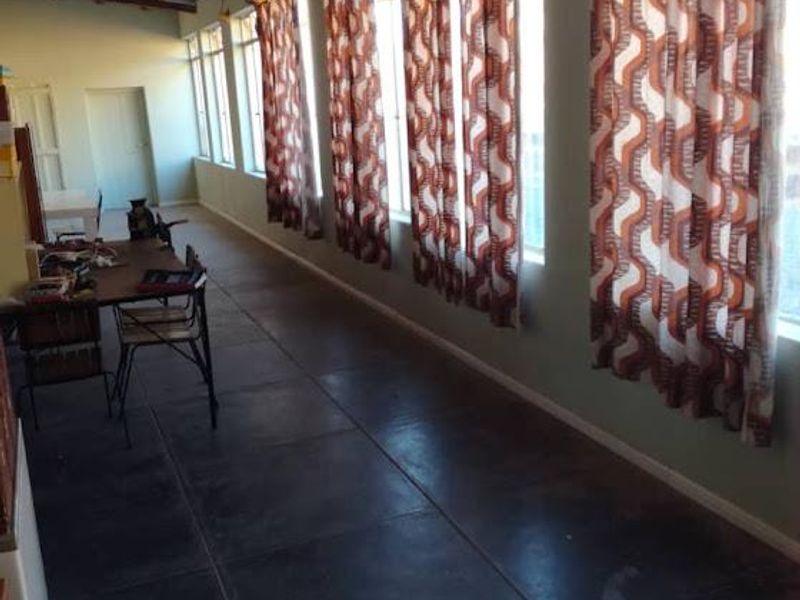 A long, sunlit enclosed patio or sunroom featuring large windows, tiled flooring, and an exposed corrugated metal ceiling.