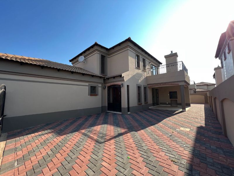 Rear exterior view of a two-story house featuring a paved driveway, a covered patio with a built-in braai, and an upper-level balcony.