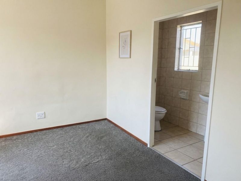 A clean, empty bedroom featuring neutral carpeting and direct access to an en-suite bathroom with tiled finishes and a window.