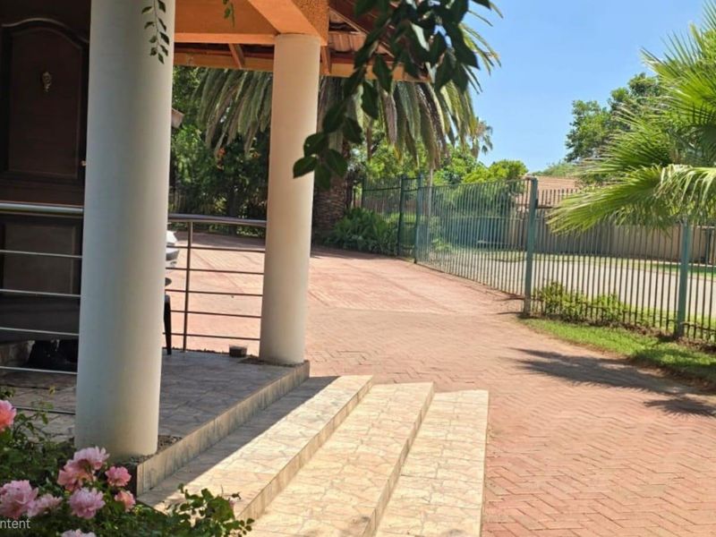 Front entrance area featuring a covered porch with concrete columns, tiled steps, and a paved driveway enclosed by metal perimeter fencing.