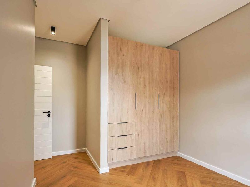 A modern bedroom featuring high-quality herringbone timber flooring, neutral wall tones, and a sleek, built-in light wood wardrobe with integrated drawers.