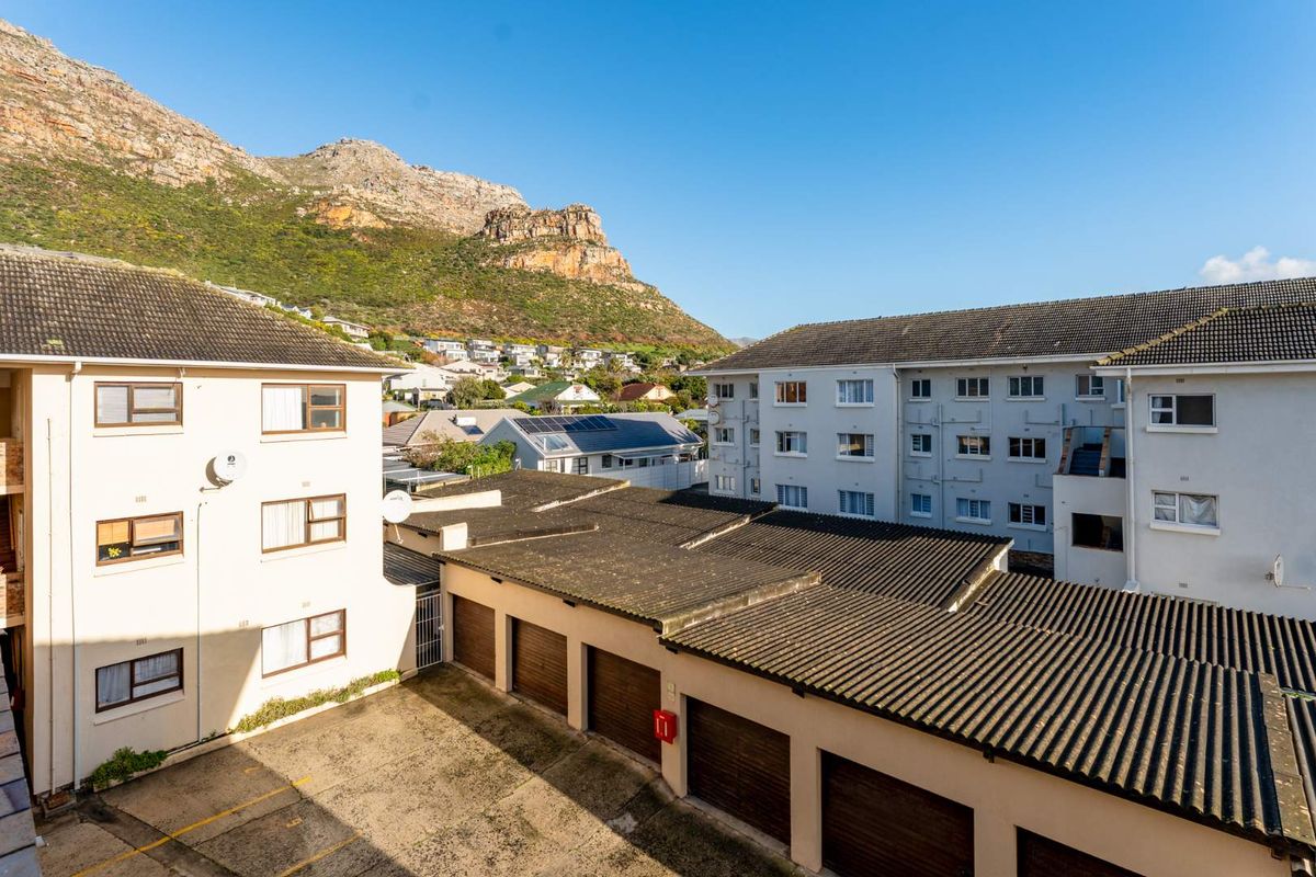 An elevated view of the apartment complex courtyard featuring a row of garages, paved parking area, and surrounding residential buildings against a mountain backdrop.