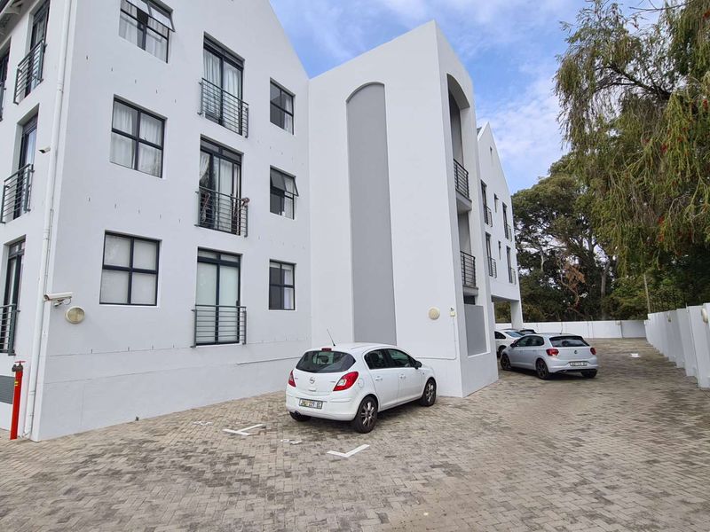 Exterior view of a modern apartment complex featuring a paved parking area, white facade, and private balconies.
