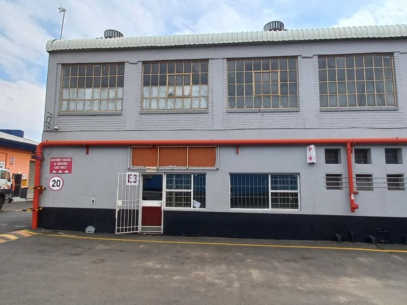 Front elevation of an industrial warehouse building featuring a security gate, steel-framed windows, and exposed exterior piping.