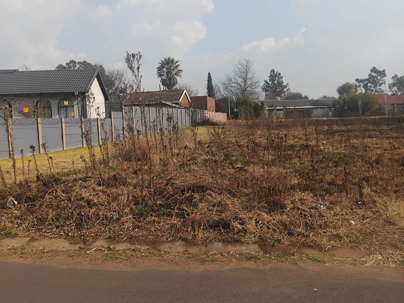 A vacant residential plot featuring a perimeter wall with security fencing, situated along a paved road in a developed neighborhood.