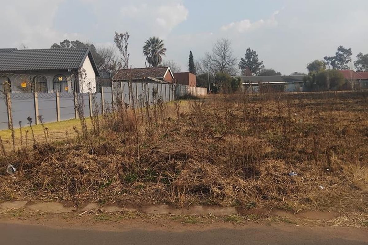 A vacant residential plot featuring a perimeter wall with security fencing, situated along a paved road in a developed neighborhood.