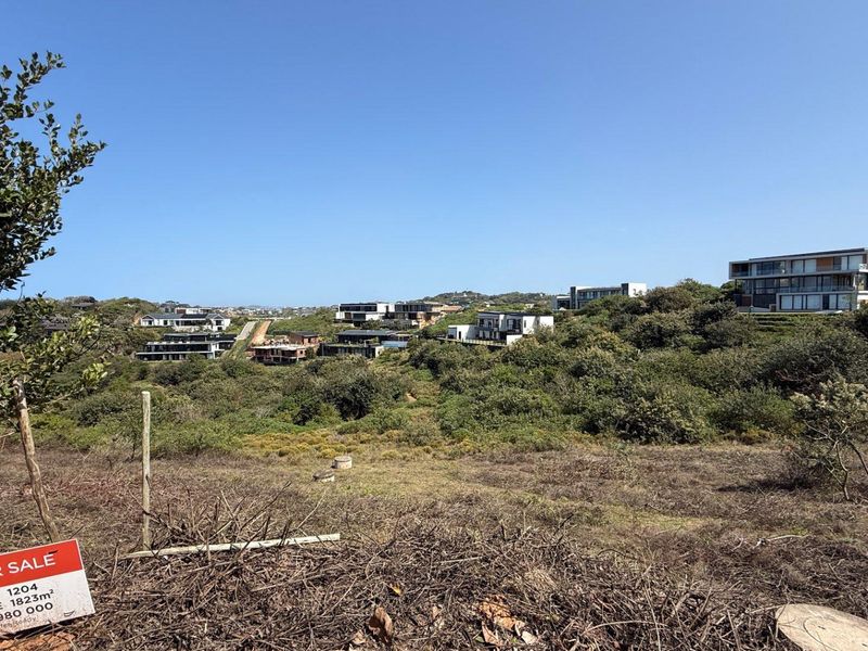 A view of a vacant residential plot featuring natural vegetation and a gentle slope, overlooking the surrounding estate and modern homes.