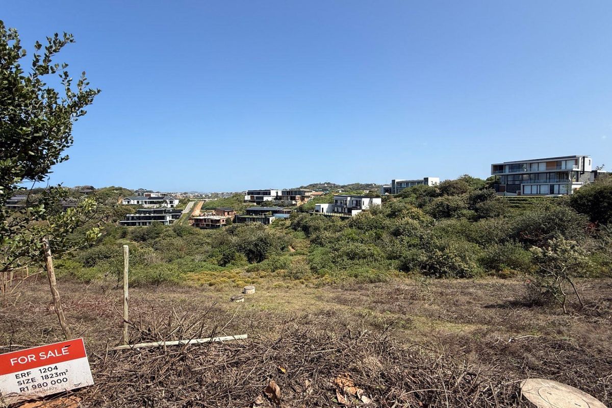 A view of a vacant residential plot featuring natural vegetation and a gentle slope, overlooking the surrounding estate and modern homes.