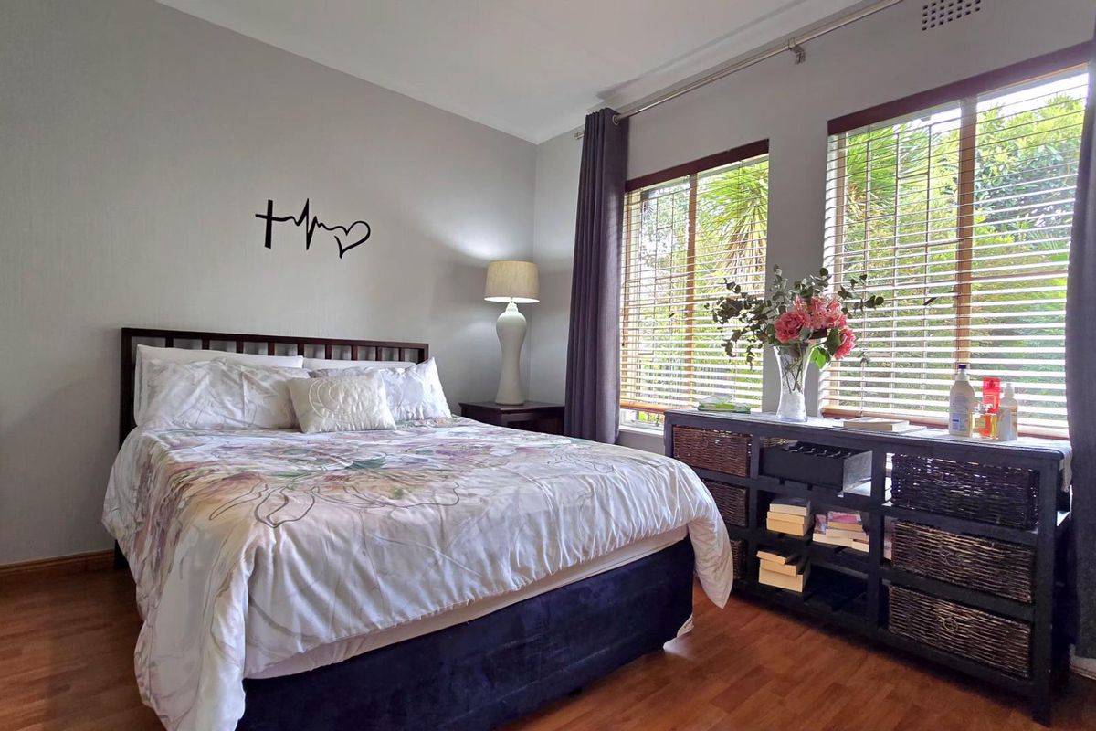 A well-lit bedroom featuring warm-toned laminate flooring, large windows with fitted blinds, and neutral wall finishes.