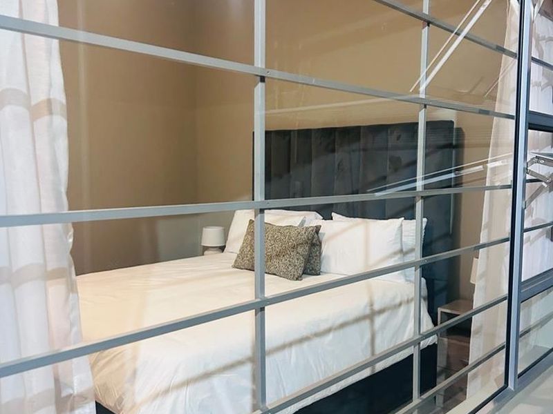 A bedroom viewed through a large window with a prominent metal grid frame, showing a neutral interior space.
