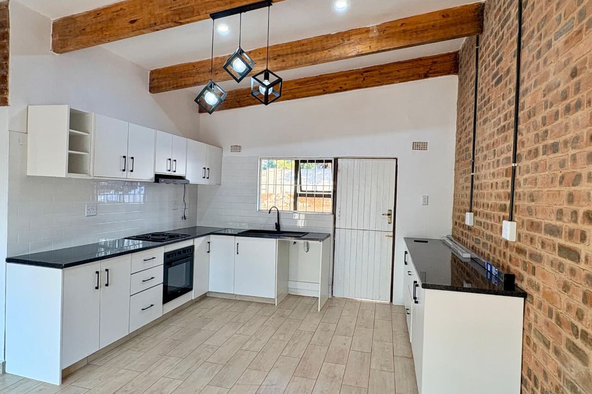 Modern kitchen featuring white cabinetry, black countertops, exposed wooden ceiling beams, and a rustic brick feature wall.