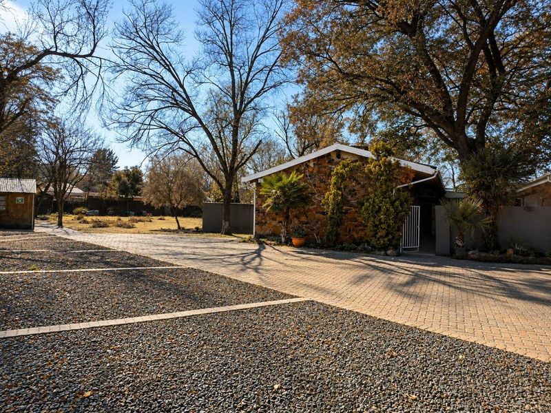 Front exterior view of the property featuring a stone-clad facade, a paved driveway, and a gravel parking area surrounded by mature trees.