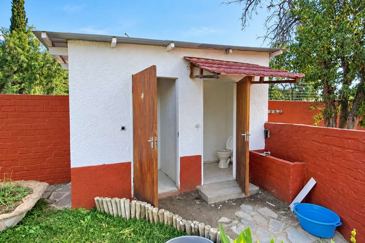 A detached outdoor utility structure featuring a toilet and basin, set within a garden area with brick boundary walls.