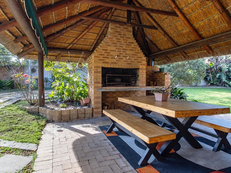 Covered outdoor patio area featuring a traditional thatched roof, a built-in brick braai, and brick paving, overlooking a landscaped garden.