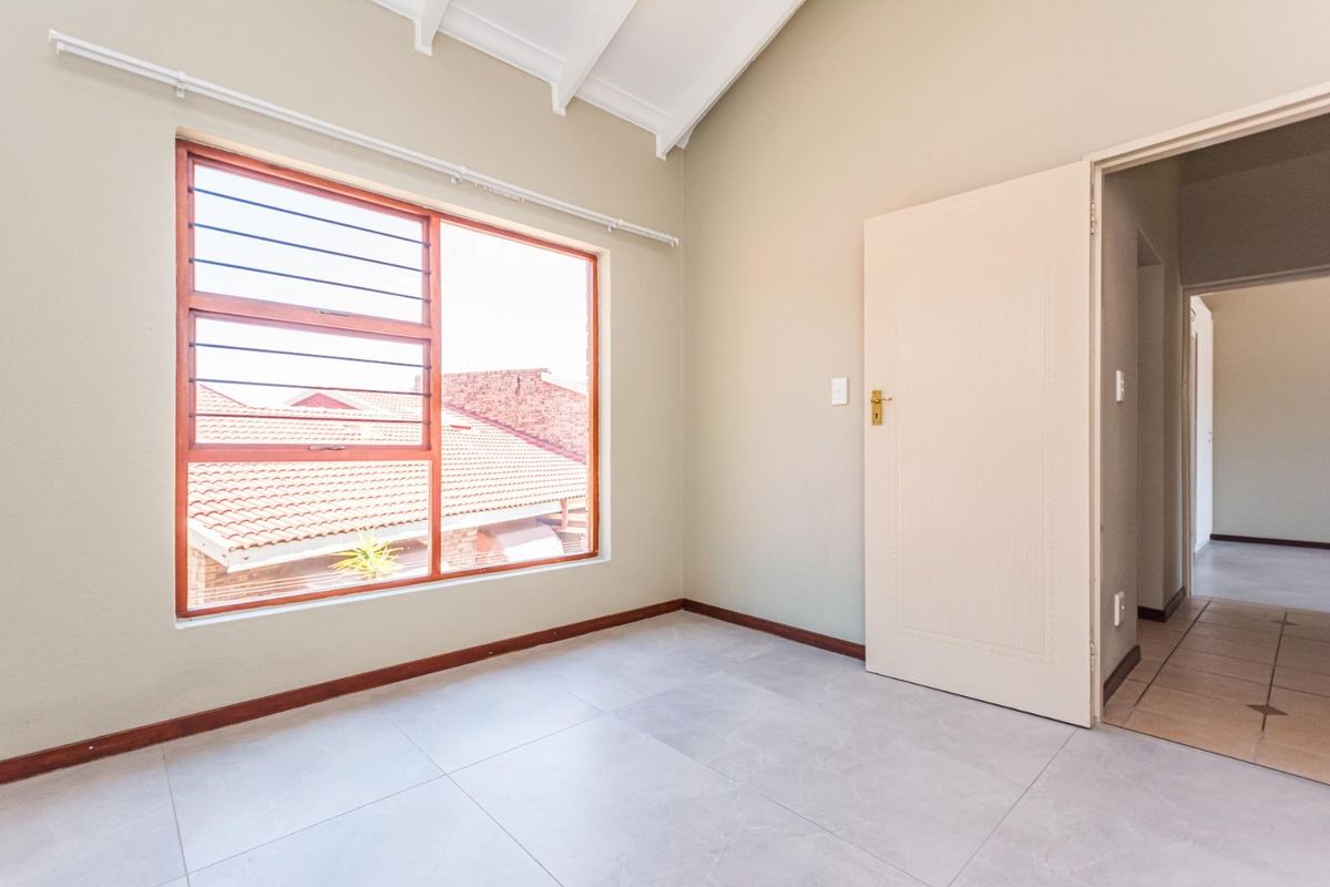 A bright, empty bedroom featuring tiled flooring, a vaulted ceiling with exposed rafters, and a large window with security bars.