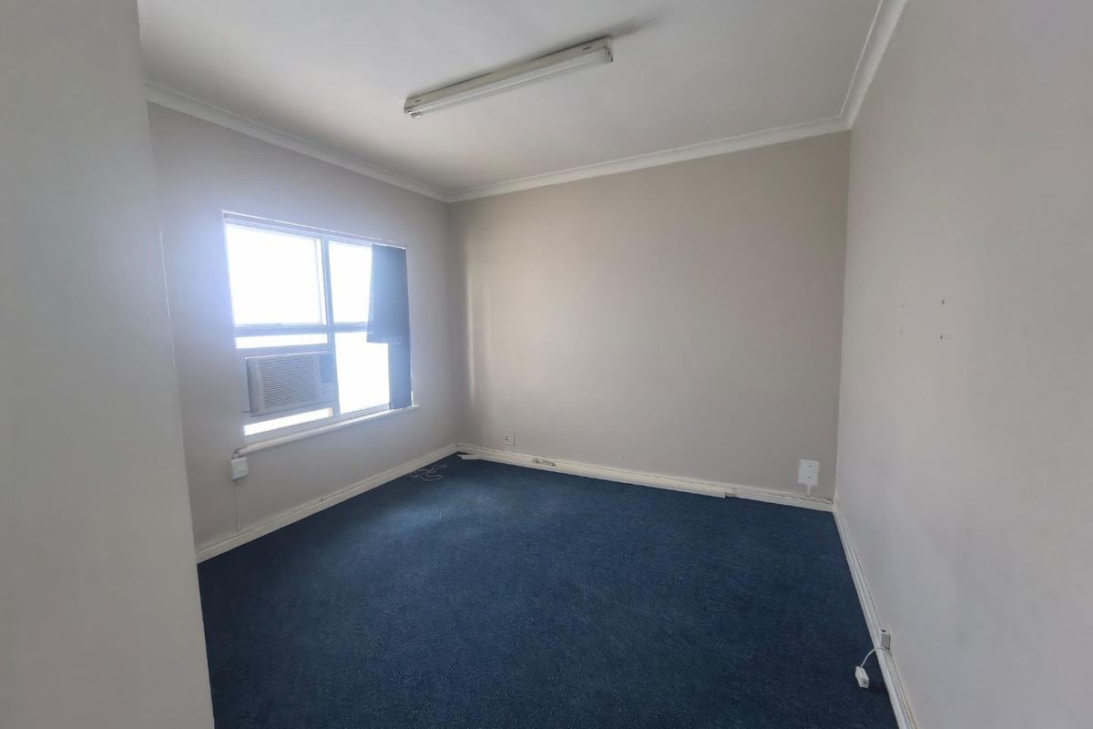 An empty office space featuring blue carpeting, a window-mounted air conditioning unit, and neutral wall finishes.