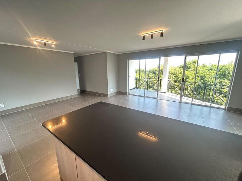 Spacious open-plan living area featuring a modern kitchen island with granite countertop, neutral tiled flooring, and large sliding doors leading to a private balcony.